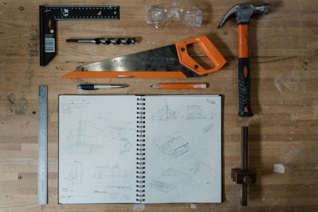 A saw, hammer, ruler, and sketchpad arranged on a workbench, displaying woodworking tools and designs.